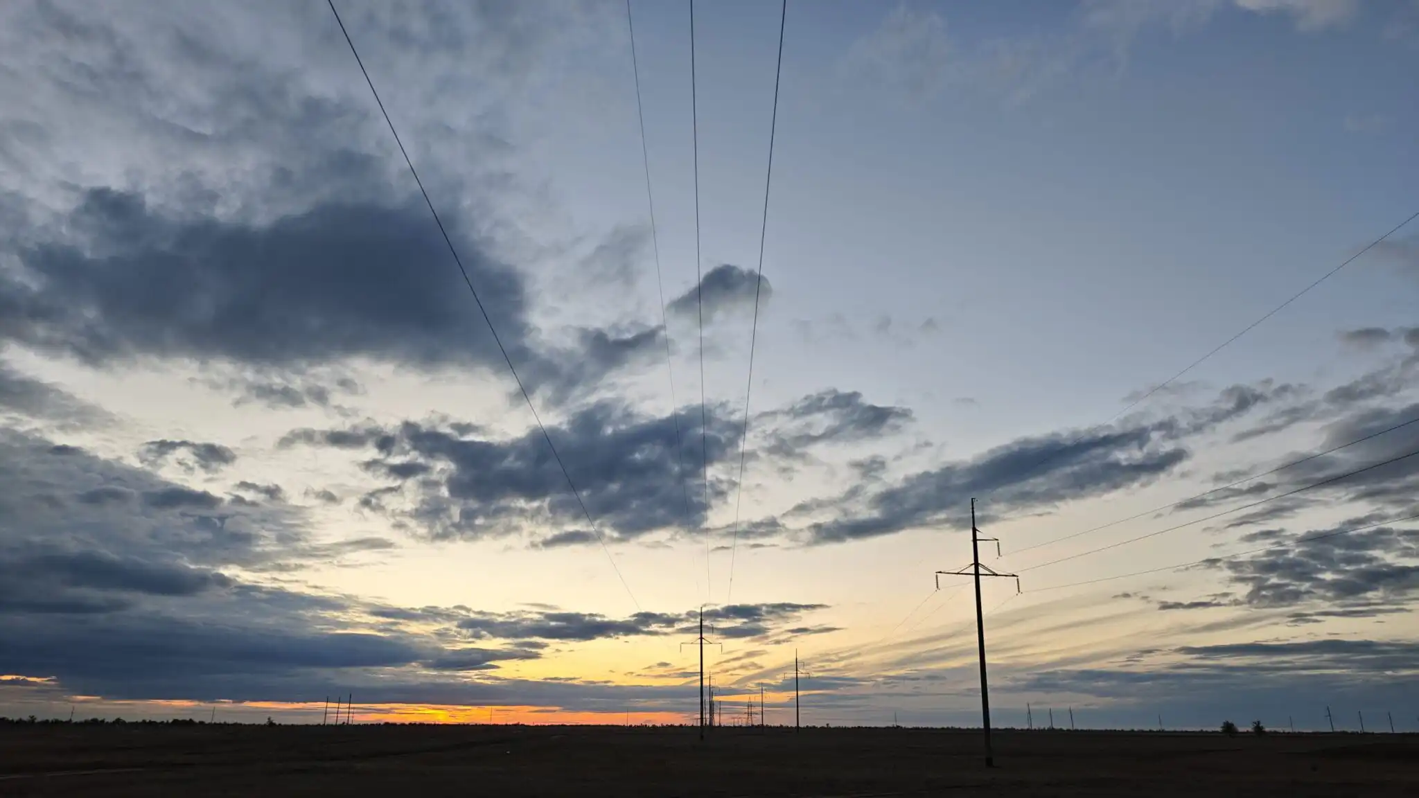 Power lines and electricity pylons stretch across a vast field under a dramatic sunset sky, Kazakhstan
