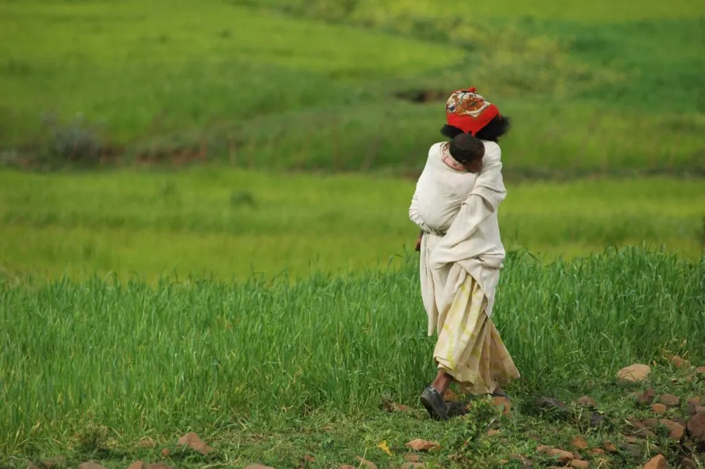 Ethiopian Woman carry baby on back in green fields.